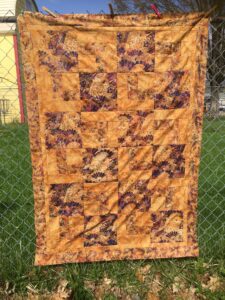 Medium brown and burgundy quilt top photographed on chain link fence with grass and yellow house in the background