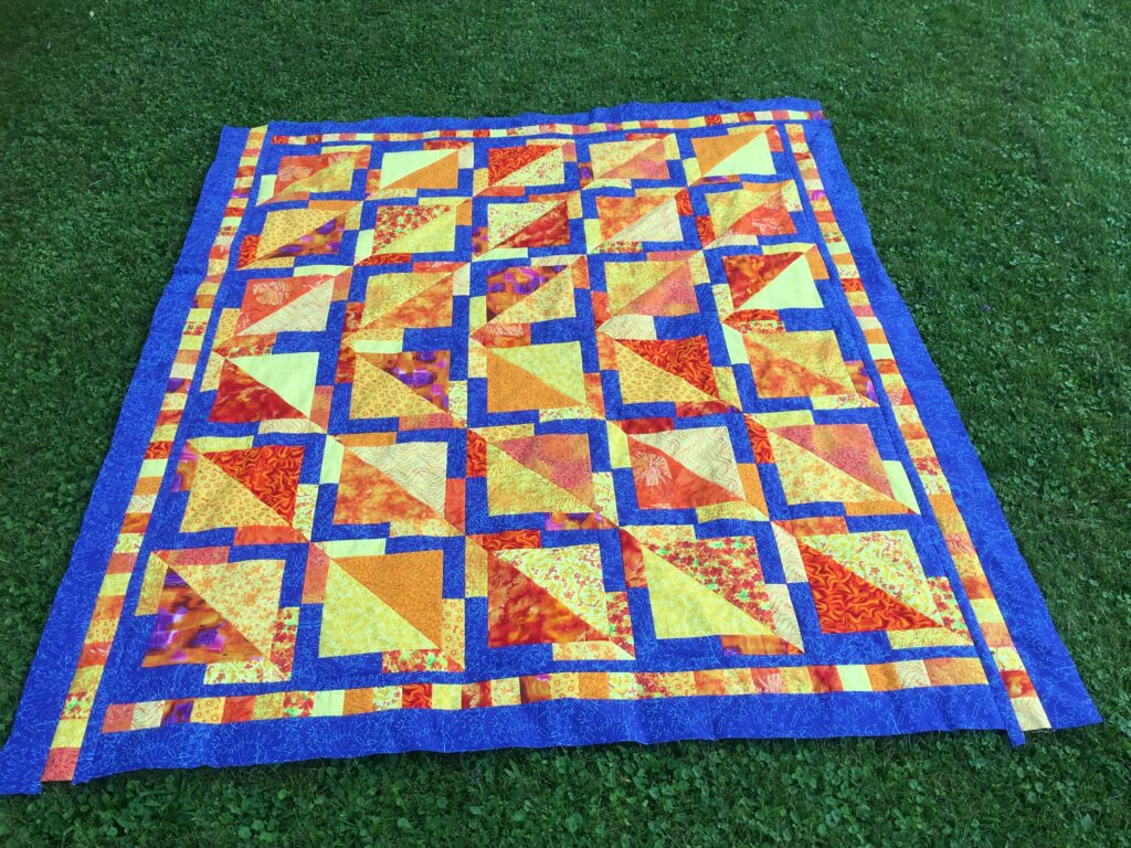 Orange and yellow quilt top with royal blue accents, on green grass.