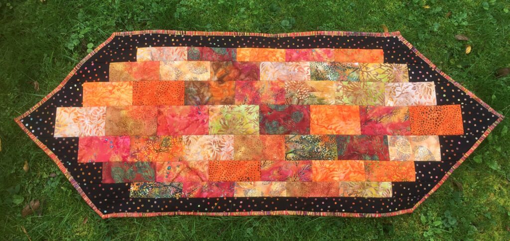 Photo of a table runner in an elongated hexagon shape. Primarily in orange, red and tan colors, with a black border and orange/green binding.